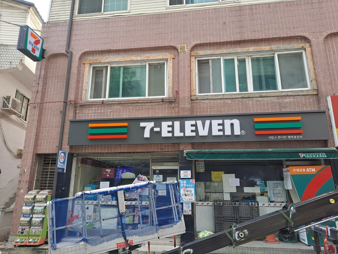 분홍 타일 건물 1층의 세븐일레븐 전면 간판과 설치 중인 고소 작업차